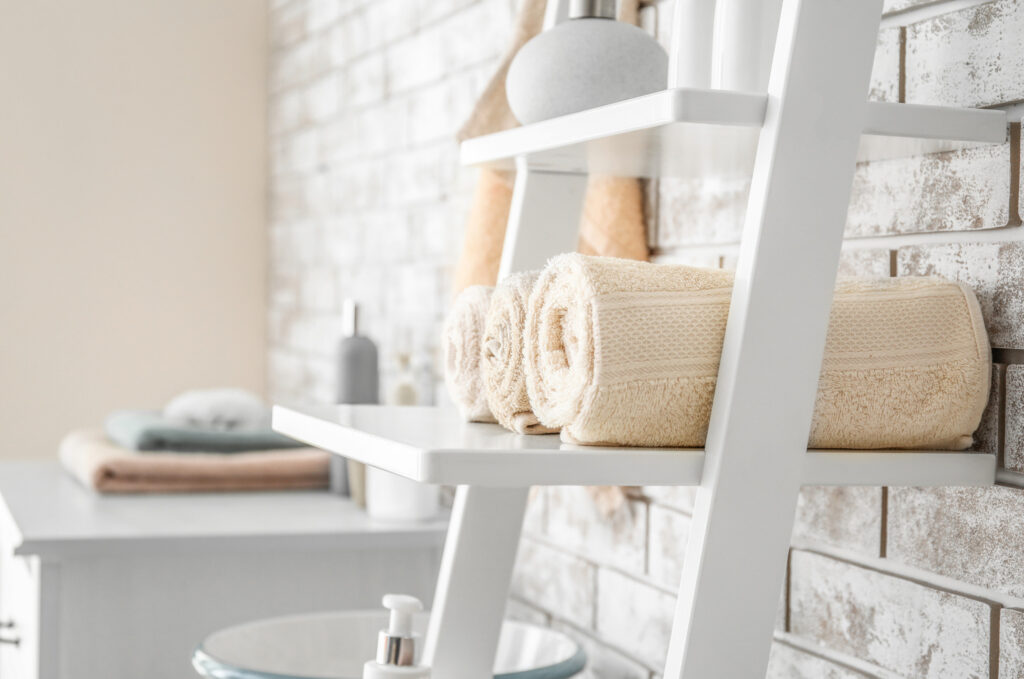 Soft beige plush towels neatly rolled on a white wooden corner shelf in a cozy bathroom.
