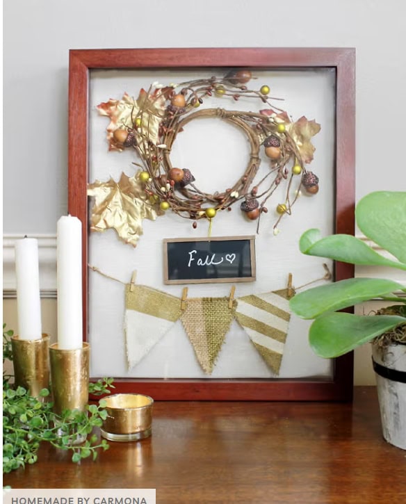 Autumn fall wreath with gold leaves, acorns, and berries displayed in a wood frame on a wall, decorated with fall-themed accents, cozy home decor, and a small chalkboard with "Fall" written on it.