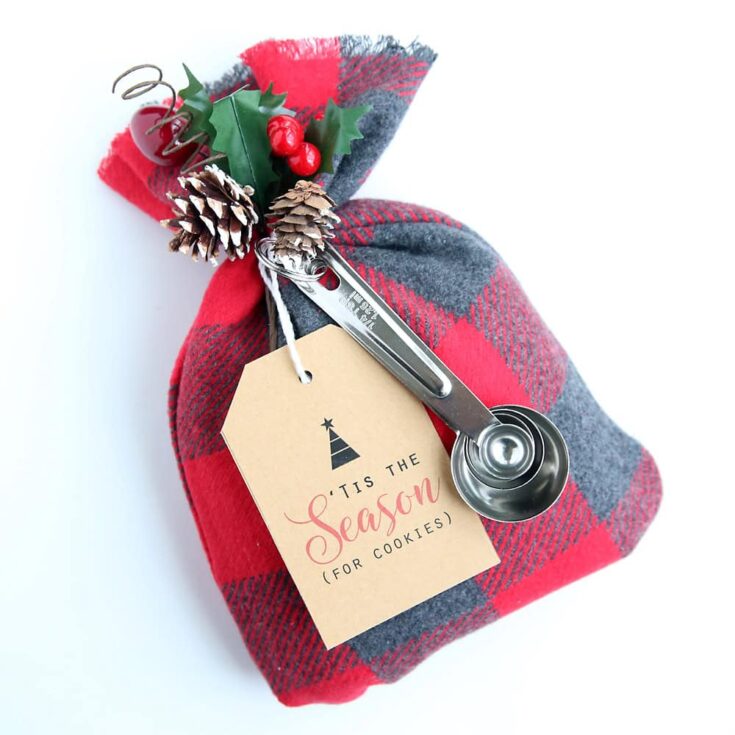 Festive holiday gift wrapped in red and gray plaid fabric with holiday décor, spoon, and a seasonal tag reading "Tis the Season for Cookies" for Christmas or winter baking traditions.