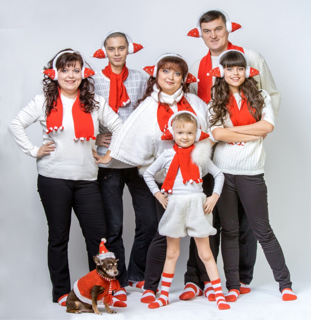 Family dressed in festive holiday pajamas and accessories, celebrating Christmas with matching socks, Santa hats, and cozy sweaters. Includes a small dog also in holiday attire, creating a joyful and warm holiday scene.