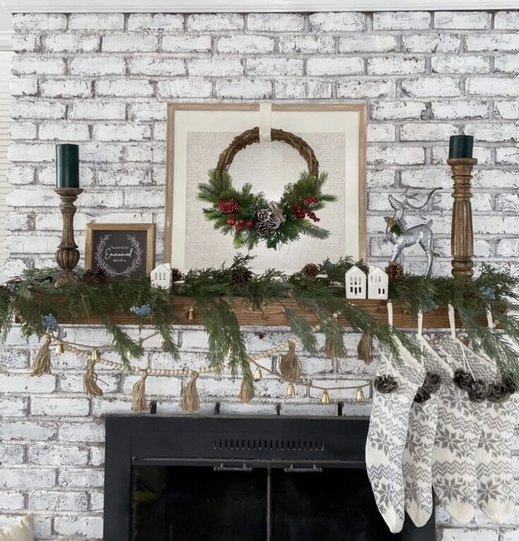 Pine wreath with red berries and pinecones hanging above a decorated fireplace mantel, surrounded by candles, small ornaments, and holiday greenery, creating a cozy Christmas decor scene.