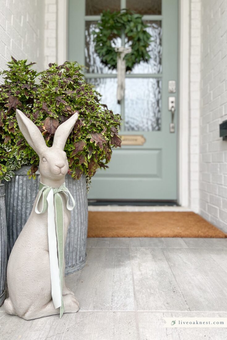 Decorative bunny statue with a bow next to lush potted plants on a cozy front porch, showcasing charming home decor and outdoor garden ideas.
