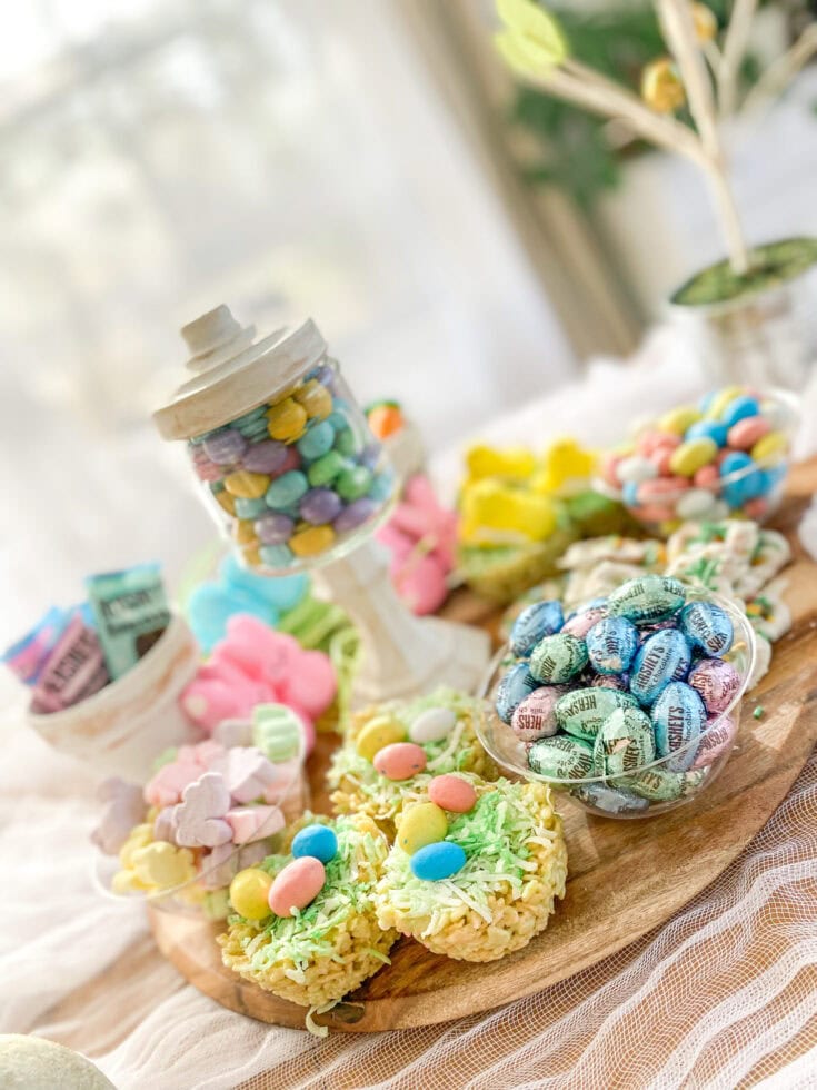 Colorful Easter candy assortment including Reese's and Jelly Belly chocolates on a wooden platter, with marshmallow treats and a potted plant in the background, perfect for spring and celebration desserts.