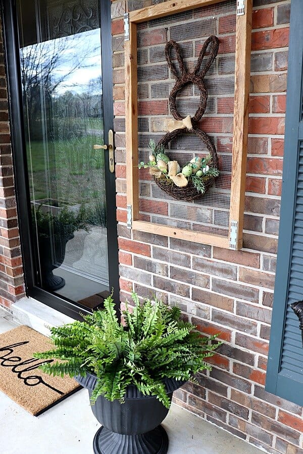 Whimsical bunny-shaped wreath with greenery and natural decor hanging on a brick wall next to a black front door.