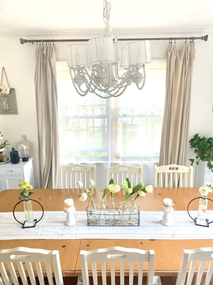 Vase with white flowers on a dining table, decorated with candles and a lace table runner, set in a bright, airy dining room with large windows and elegant curtains.