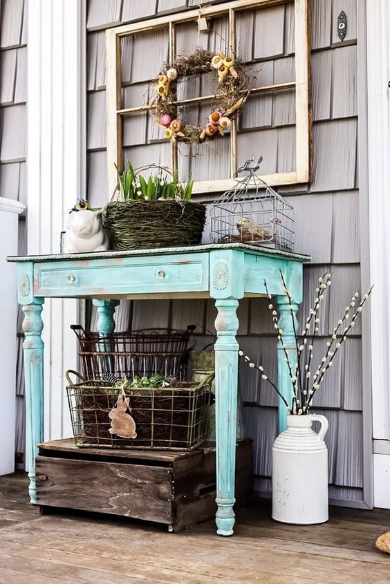 Rustic outdoor decor with a distressed turquoise table, spring flowers in a white pitcher, and decorative accents for farmhouse-style porch or garden entry.