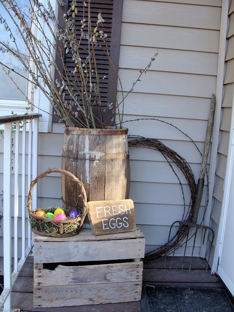 Rustic outdoor spring decor with a vintage wooden barrel, Easter eggs basket, and a "Fresh Eggs" sign on a porch.