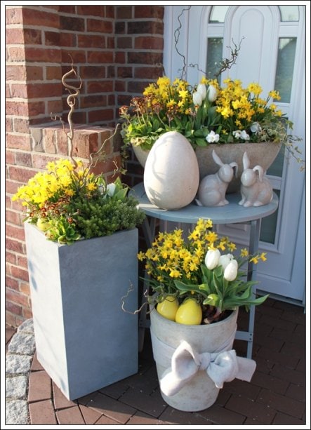 Bright spring flower arrangements with yellow, white, and green blooms, decorative Easter eggs, and bunny accents displayed outdoors near a brick wall and white door.