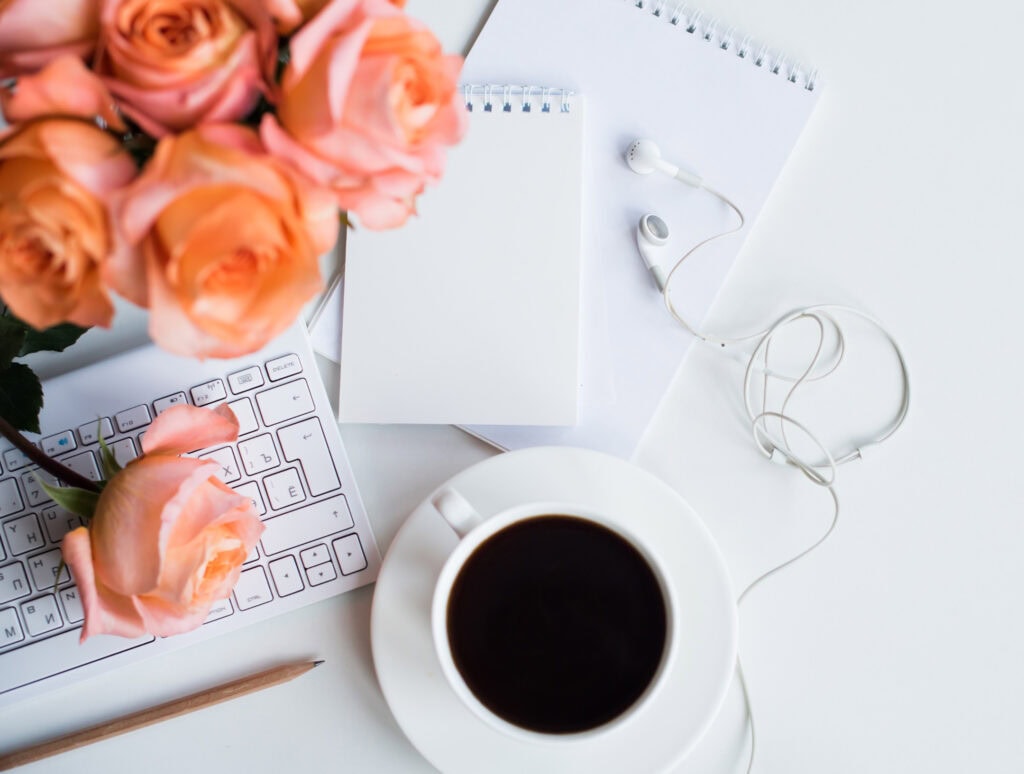 Soft pink roses on a clean workspace with a keyboard, notepad, coffee, and earphones, perfect for creating relatable blog content or lifestyle photographs for A Hundred Affections.