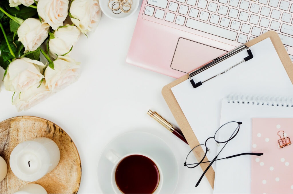 Delicate white roses with green leaves on a white workspace with a pink laptop, notebooks, glasses, and coffee, perfect for a romantic or feminine lifestyle blog.