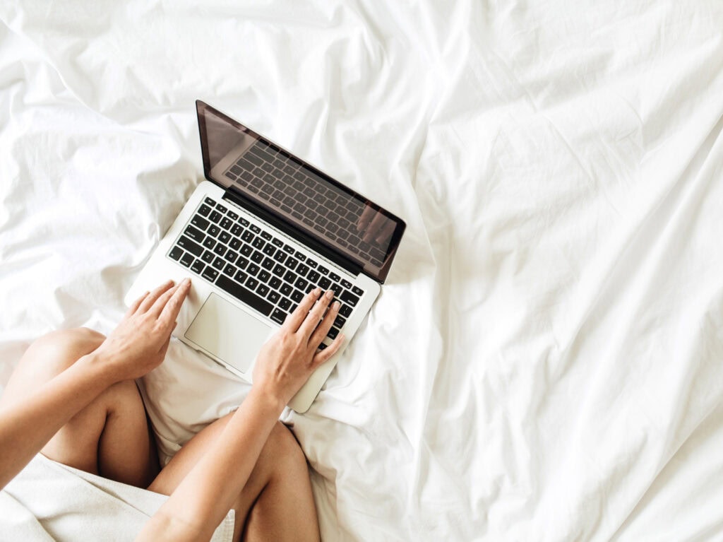 Laptop on bed with woman typing, cozy bedroom workspace, modern device for remote work, comfortable home office, digital technology in a relaxed setting.
