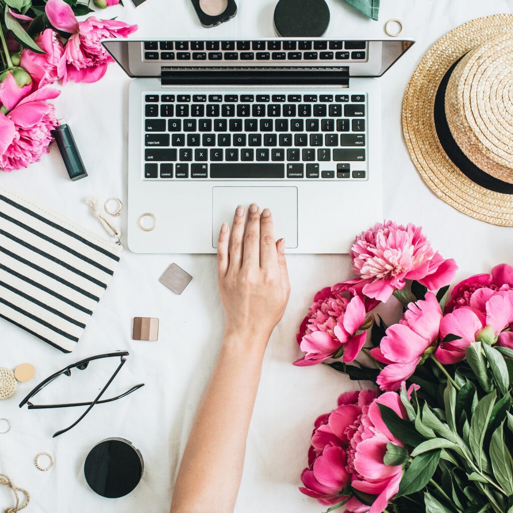 Vibrant pink peony flowers surrounding a silver laptop on a white desk with beauty and fashion accessories.