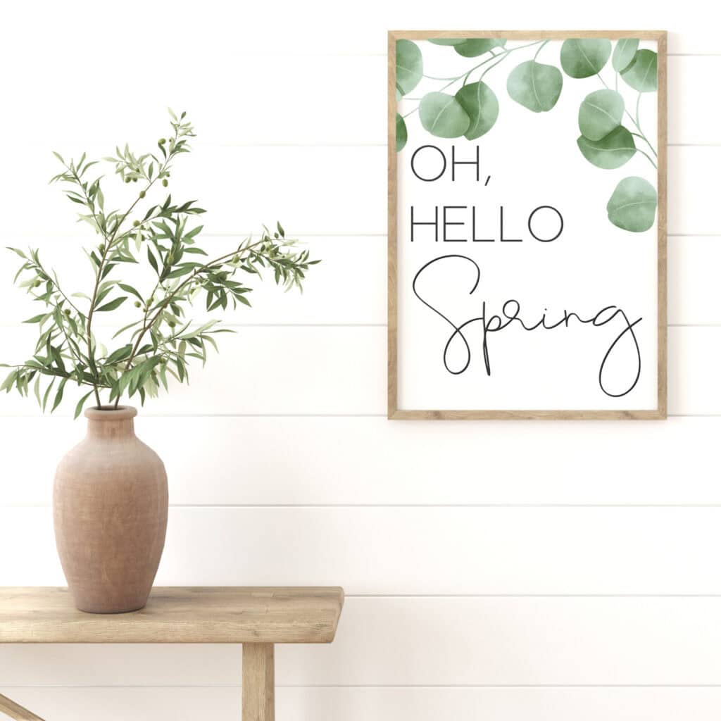 Green eucalyptus leaves in a beige vase on a wooden table, with a spring-themed wall art sign that reads "Oh, Hello Spring" in stylish calligraphy.