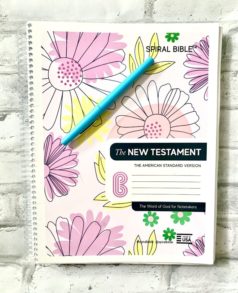 Aspirational floral-themed spiral Bible journal with colorful pink, yellow, and green illustrations, featuring the New Testament and a blue pen resting on the pages for note-taking and journaling.
