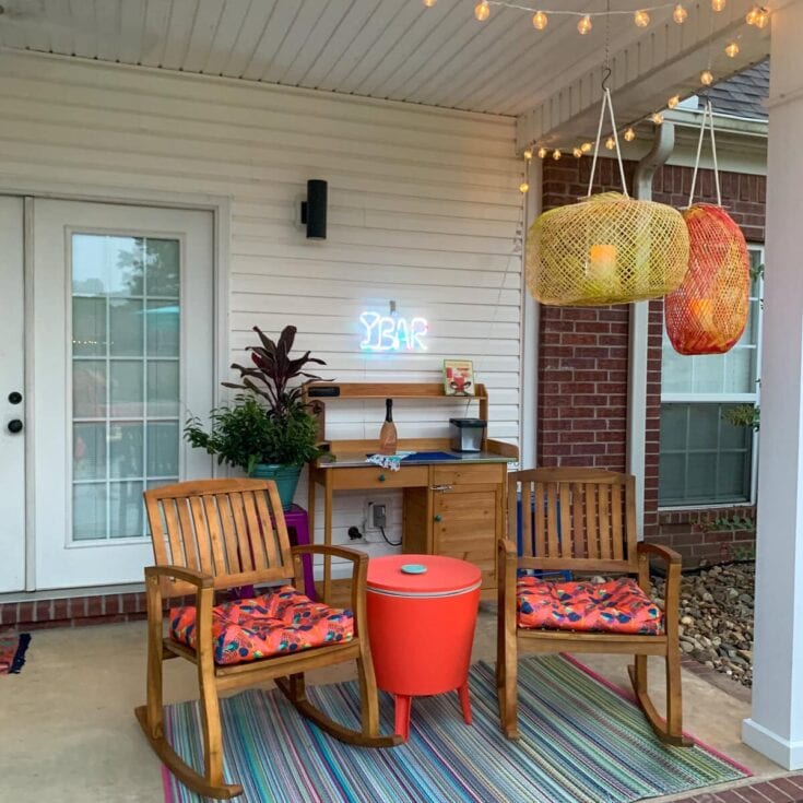Charming outdoor patio setup featuring wooden rocking chairs with colorful cushions, hanging lanterns, string lights, and a cozy bar area against a white siding wall, perfect for relaxing and entertaining.