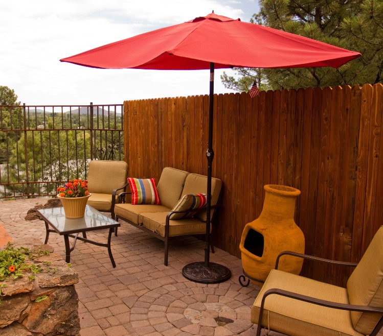 Cozy outdoor patio with seating, a large red umbrella, decorative pillows, and garden accents perfect for relaxing and enjoying nature.