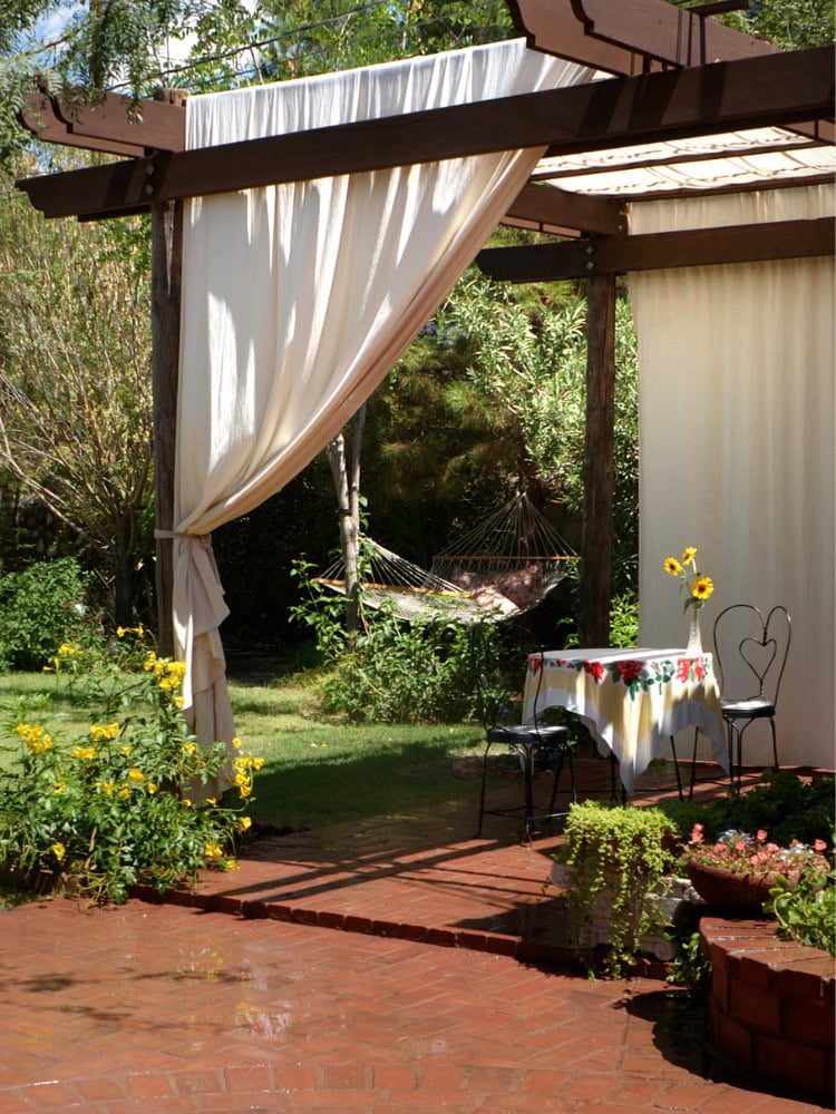 Charming outdoor patio with beige curtains, vintage table with sunflower centerpiece, lush garden background, perfect for romantic gatherings or relaxing outdoor retreats.