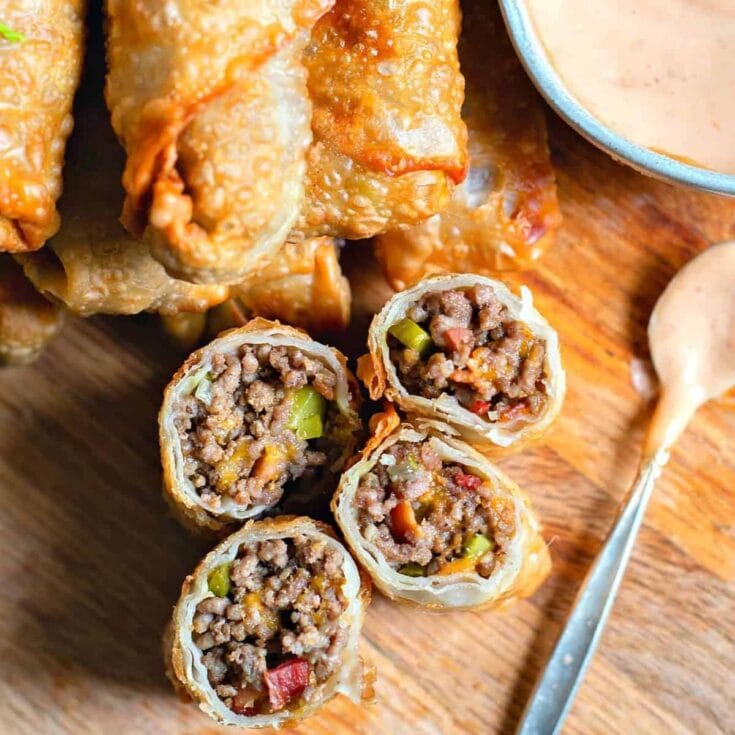 Hearty beef and vegetable egg rolls with crispy golden exterior, sliced open to reveal savory filling, served with creamy dipping sauce on a rustic wooden platter.