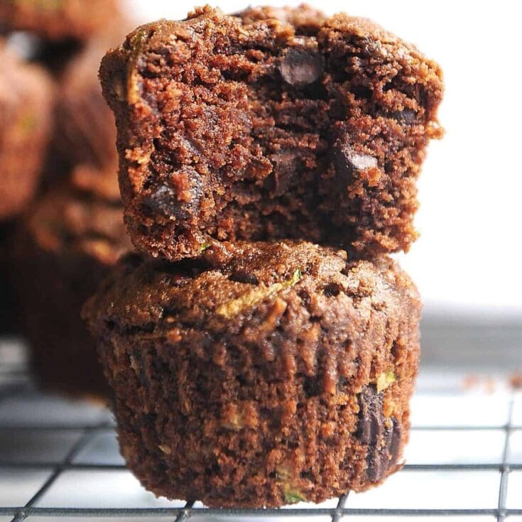 Crispy, gooey chocolate zucchini muffins stacked on a wire rack showing moist interior with chocolate chunks. Perfect for healthy baking, featuring zucchini and rich chocolate flavor.