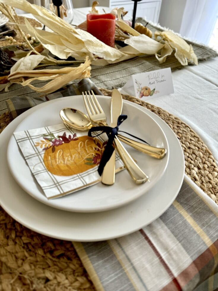 Gold cutlery set on a white plate with a autumn-themed napkin; elegant Thanksgiving table setting with fall decor and a "thankful" sentiment.