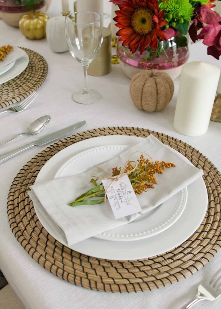 A beautifully set table with a white plate on a woven rattan placemat, decorated with a napkin wrapped with a small bouquet of yellow and green flowers, perfect for a rustic or boho-themed event.