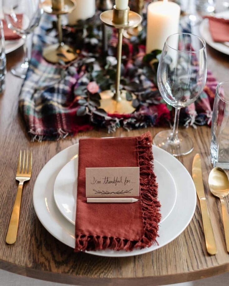 Elegant table setting with gold flatware, a white plate, and a burnt orange cloth napkin with a handwritten thankful note, surrounded by candles and greenery for a warm, inviting atmosphere.