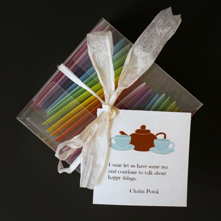 Colorful tea bag tags in a clear plastic box with a handwritten quote about enjoying tea and happy conversations, perfect for tea lovers and gifting.