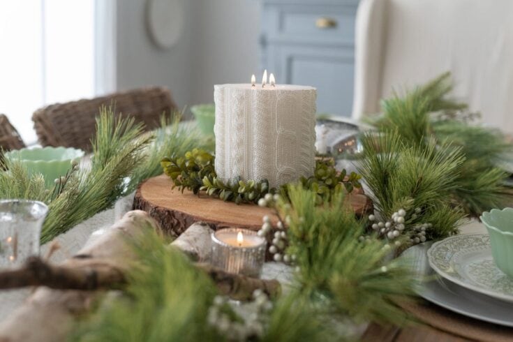 A beautifully arranged holiday table featuring a textured white candle surrounded by pine branches, berries, and wood accents for festive home decor.