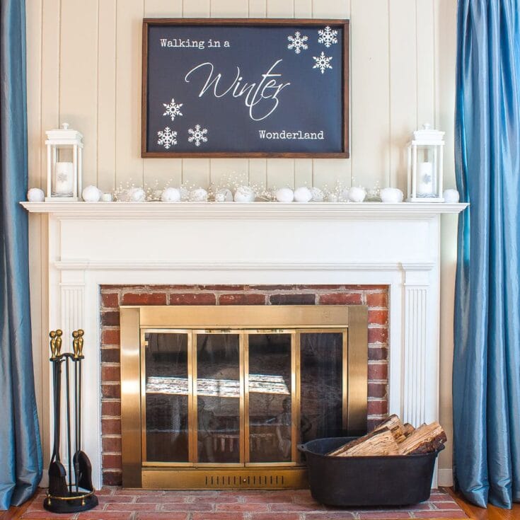 Cozy fireplace with white mantel, festive winter decor including snowballs and string lights, blue curtains, and a chalkboard sign with snowflake illustrations, perfect for a holiday winter wonderland setting.