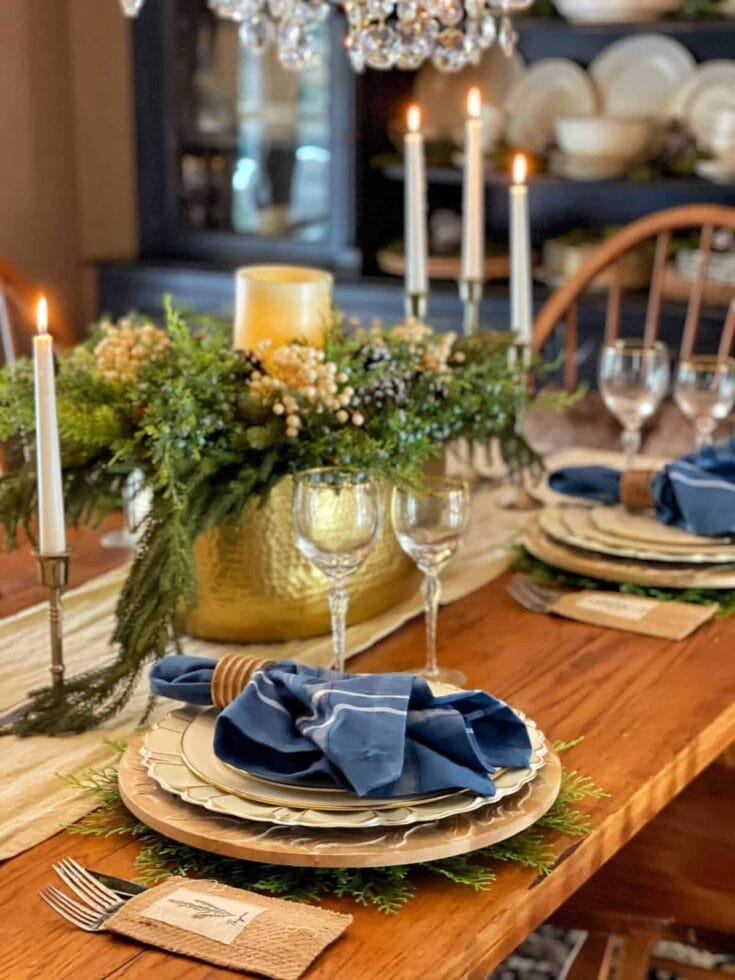 A beautifully arranged dining table with a lush greenery centerpiece, candlelit candles, and sophisticated place settings including blue napkins and gold accent plates, perfect for festive gatherings.