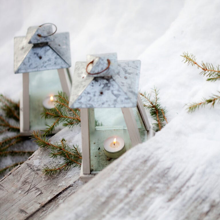 Softly glowing candle lanterns with marble and wood accents for cozy winter decor, accented with evergreen branches, creating a warm and inviting holiday atmosphere.