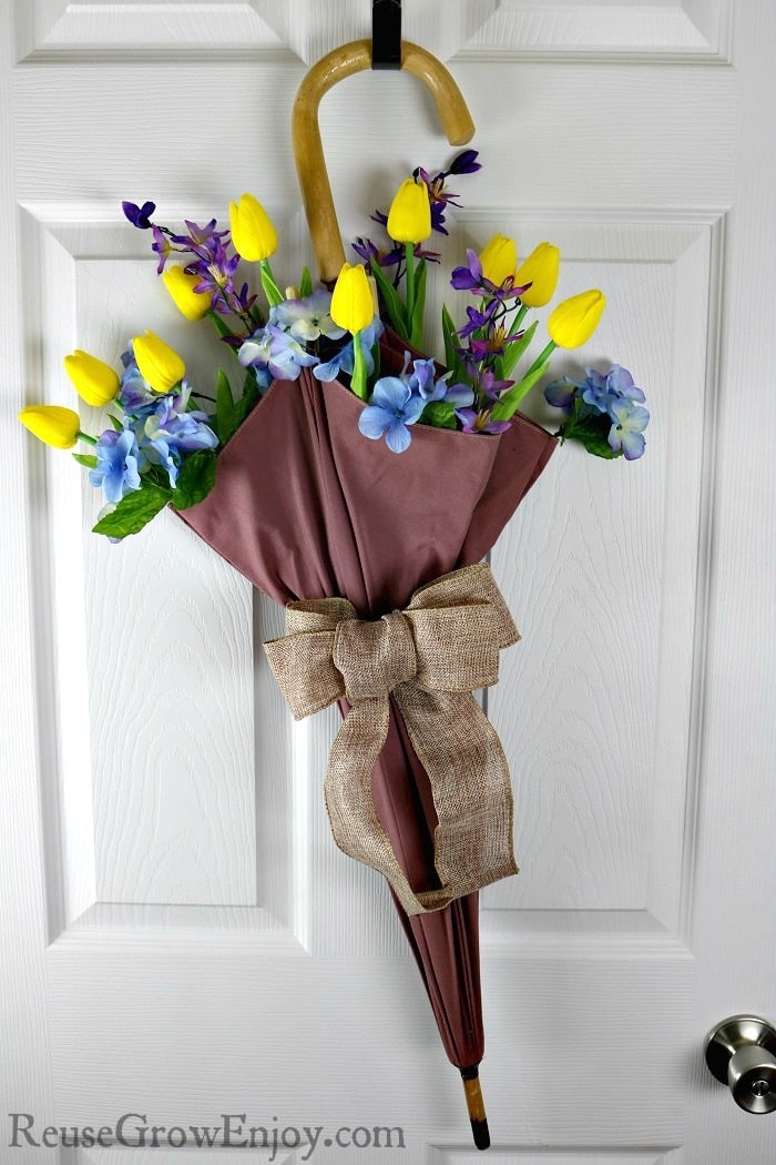 Yellow tulips, purple irises, and blue hydrangeas arranged in a floral umbrella decoration with a burlap bow on a white door.