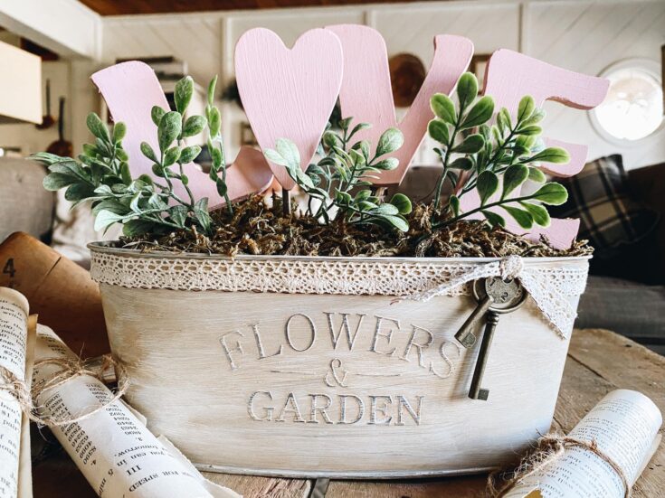Pink heart-shaped and rustic paper flower decorations in a vintage "Flowers & Garden" white wooden container with greenery and keys for a charming floral and garden-themed arrangement.
