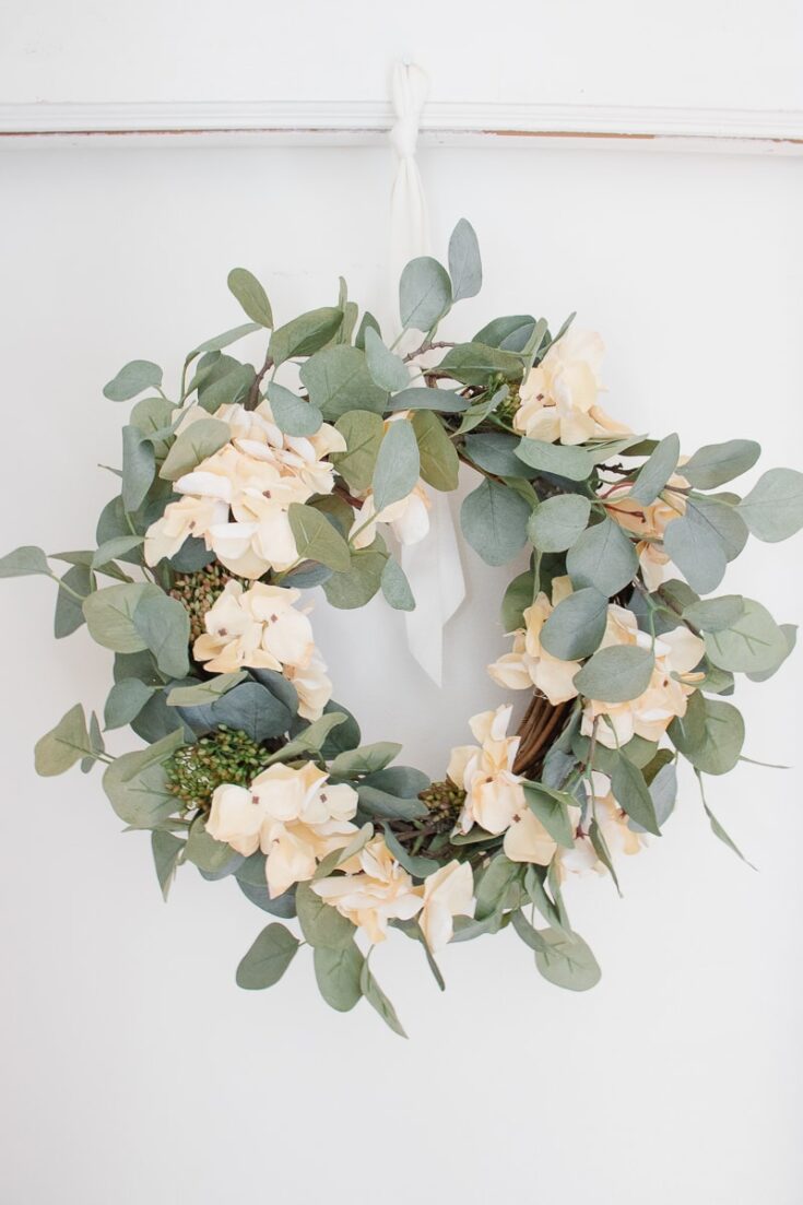 Gentle eucalyptus and cream hydrangea flower wreath hanging on a white wall, perfect for spring home decor or wedding floral arrangements.