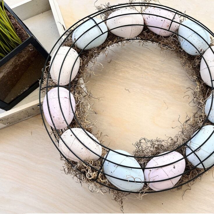 Easter egg wreath with pastel eggs on a black wire frame, nest-like decoration for spring and Easter celebrations, arranged with natural nest material on a light wooden surface.