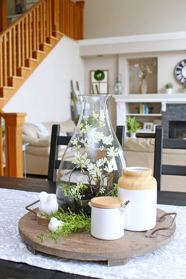 Delicate glass vase with white flowering plant, rustic wooden tray with ceramic jars, cozy living room interior with fireplace and staircase in background, home decor inspiration.