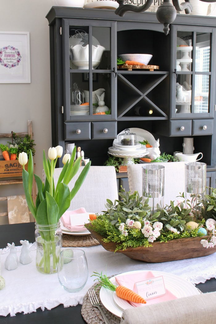 Vase with white tulips decorating a spring Easter table setting, featuring pastel-colored eggs and floral accents.