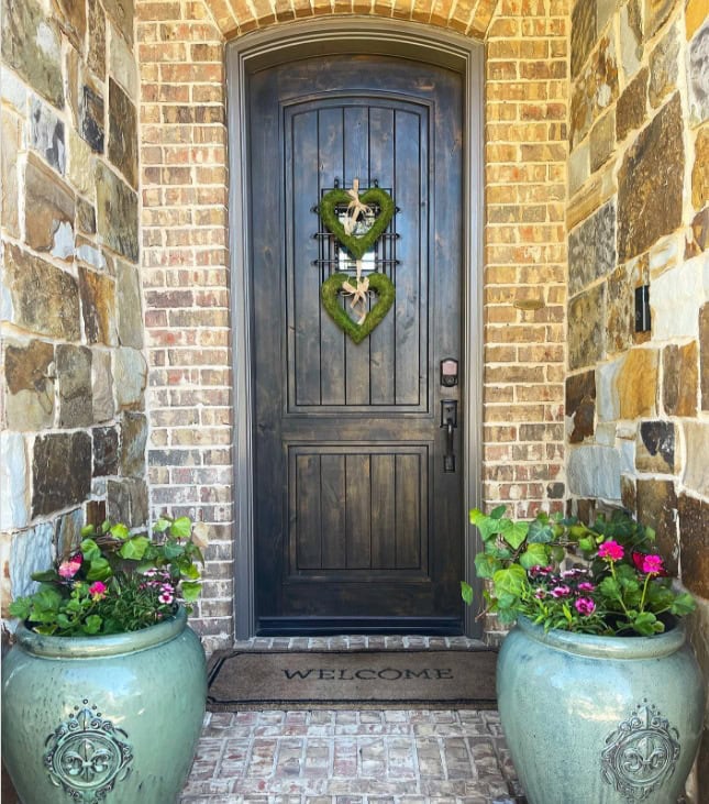 Heart-shaped wreaths with bows hanging on a dark wooden front door of a brick house, flanked by large ceramic planters filled with vibrant pink and purple flowers, creating a welcoming and charming entryway.