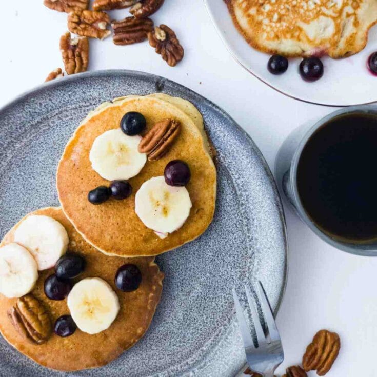 Fluffy banana pancakes topped with blueberries, banana slices, and pecans served on a textured gray plate for a delicious breakfast.