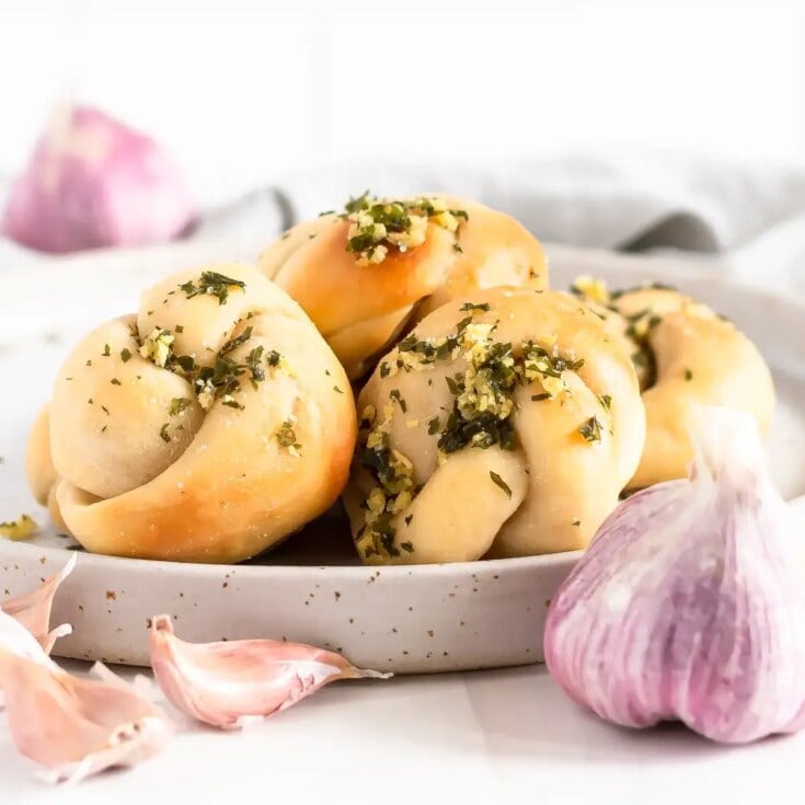 Soft garlic knots topped with chopped herbs and garlic, served on a white plate with garlic cloves and a blurred background, highlighting fresh, homemade bakery delights.