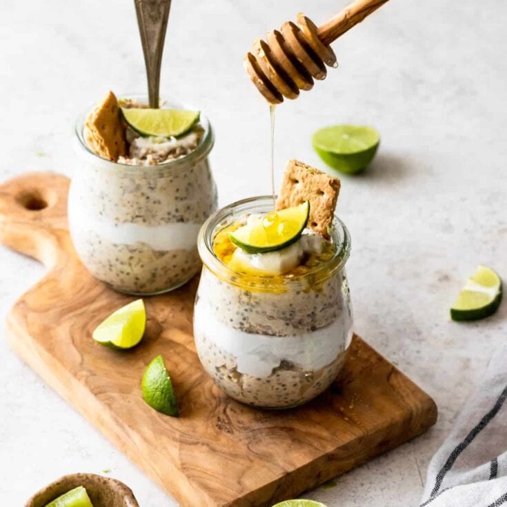 Creamy key lime pie parfaits in glass jars topped with lime wedges, honey drizzle, and graham cracker pieces, served on a wooden board with fresh lime slices for a refreshing dessert.