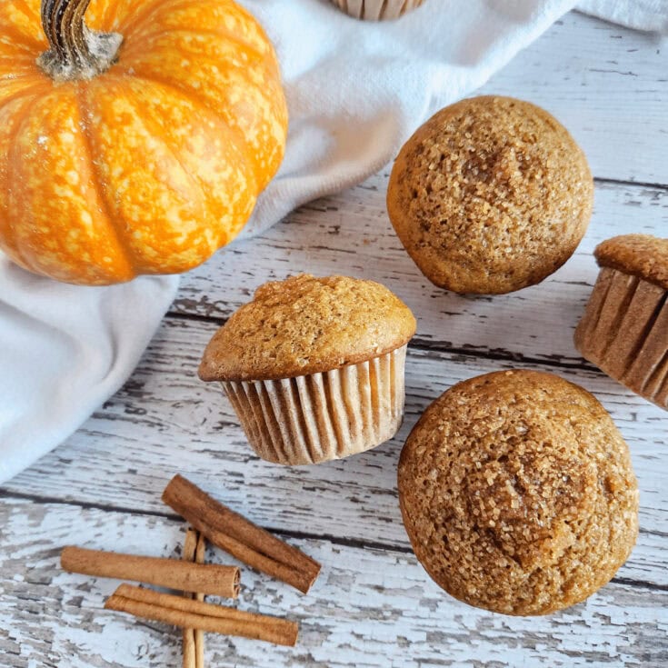 Freshly baked pumpkin muffins with cinnamon sticks and a small pumpkin on a white wooden surface, perfect for fall baking and seasonal treats.