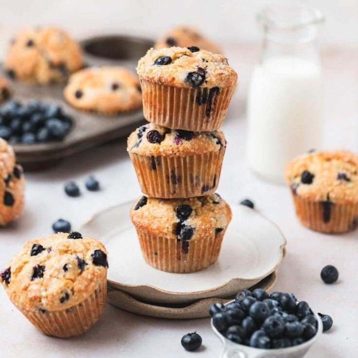 Blueberry muffins stacked on a white plate with fresh blueberries scattered around, featuring golden-brown tops and blueberry pieces throughout. Perfect for breakfast or snack, showcasing homemade berry muffin recipes.
