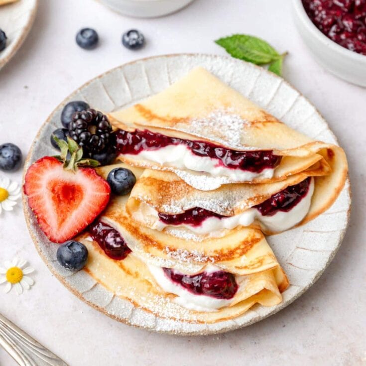 Fluffy crepes filled with mixed berries and cream, garnished with fresh strawberries and blueberries, perfect for a sweet breakfast or brunch treat.