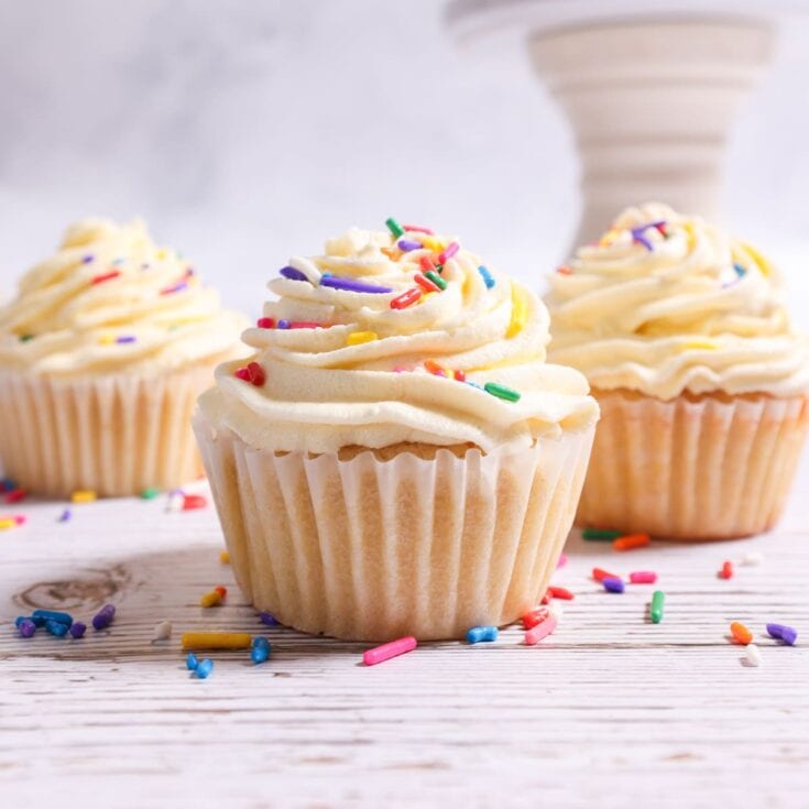 Sweet vanilla cupcakes with colorful sprinkles, topped with creamy frosting, arranged on a white wooden surface for a cheerful celebration.