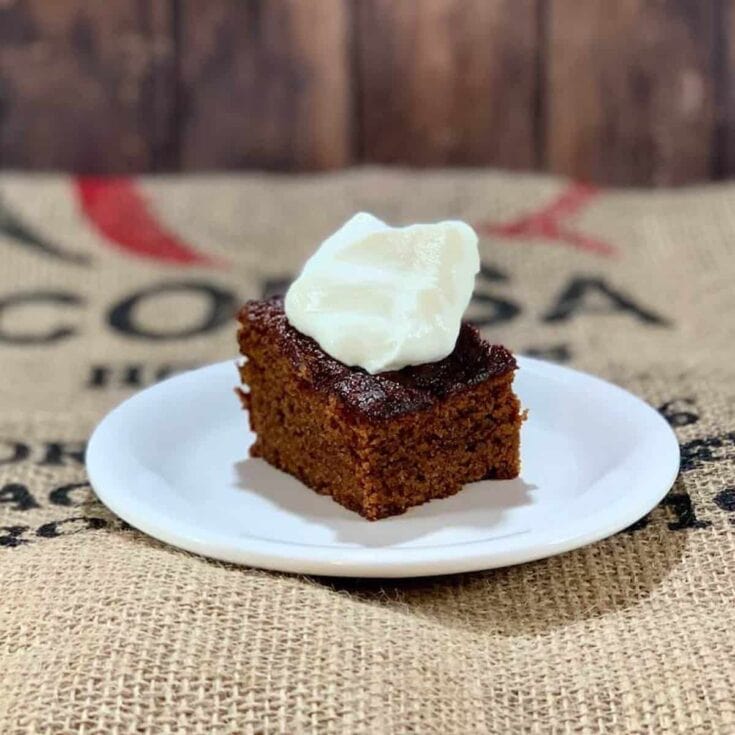 Rich moist carrot cake square topped with creamy white frosting, served on a white plate with a rustic coffee sack background for cozy, homemade dessert vibe.