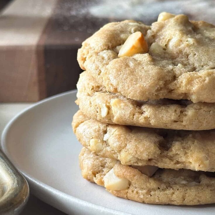 Buttery white chocolate macadamia nut cookies stacked on a white plate with a blurred background of a gift box and a jar, highlighting fresh homemade cookies, perfect for gifting or enjoying as a sweet treat.