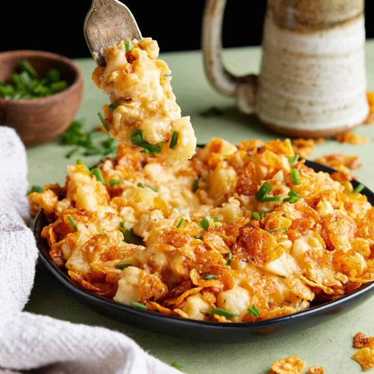 Creamy baked cheesy chicken casserole with melted cheese, crispy topping, and fresh green herbs, served in a black dish with a rustic coffee mug and bowl of chopped green onions in the background.