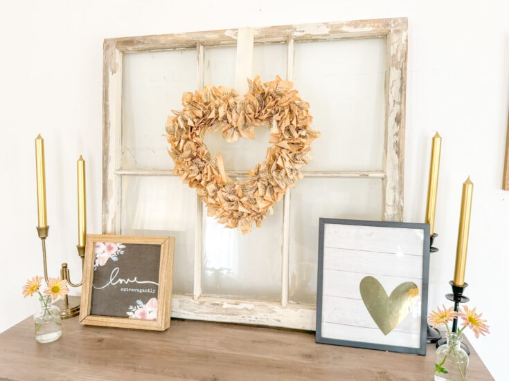 Rustic heart-shaped wreath made from crumpled pages of vintage books, displayed on an antique window frame, with decorative framed signs and small glass vases of pink daisies creates a romantic, vintage-inspired decor scene.