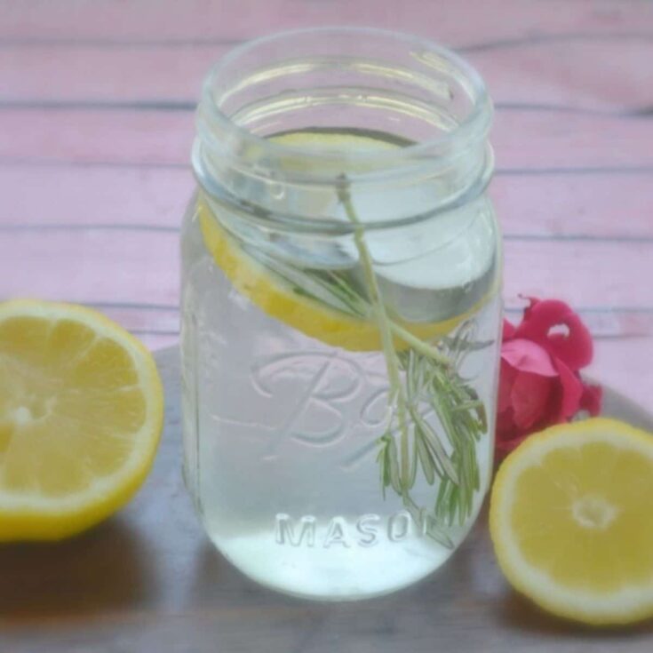 Lemon infused water in a mason jar with fresh lemon slices and rosemary, garnished with pink flowers, on a pink wooden surface, promoting healthy hydration and wellness.